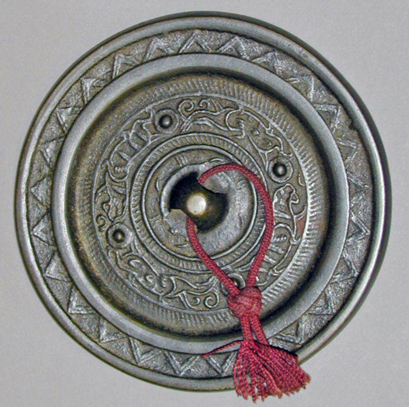 A circular bronze mirror from ancient Japan featuring intricate geometric patterns, a central knob with a red decorative cord, and raised outer rims